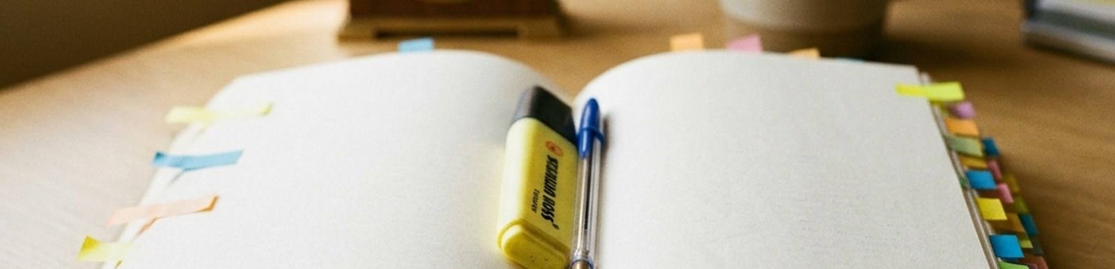 An open blank notebook with coloured index tabs and sticky flags, a highlighter and pen resting across the pages